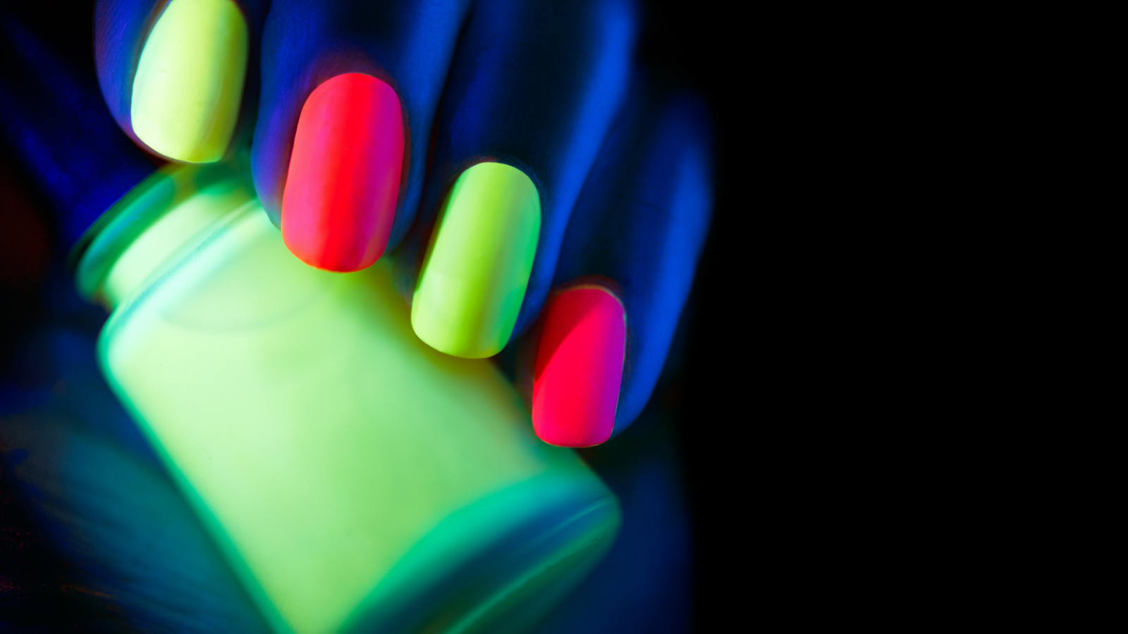 Part of a hand shown holding a bottle of fluorescent yellow nail vanish. The nails are painted with fluorescent vanish in yellow and pink.